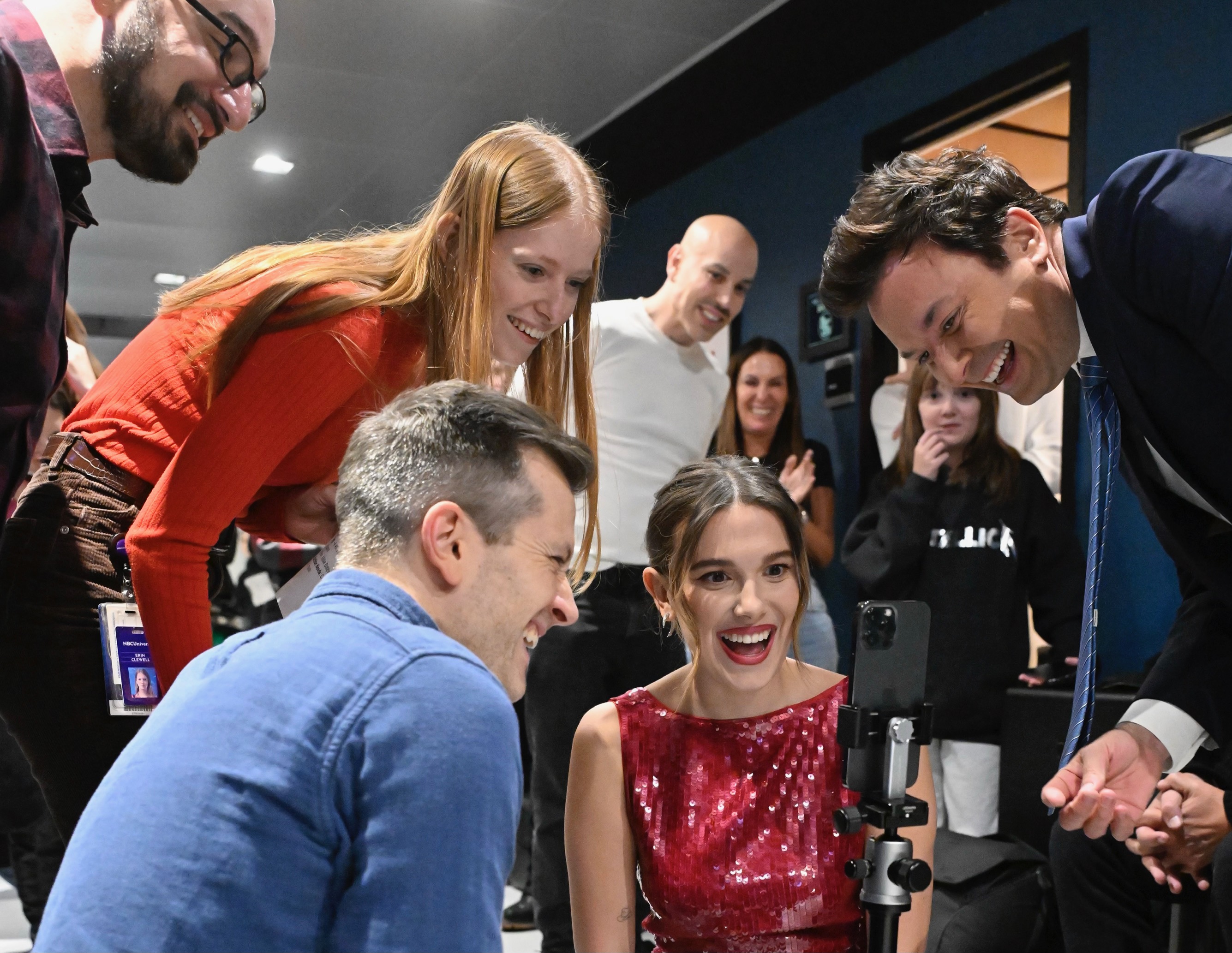 Millie Bobby Brown, wearing a red sequined dress, watches a smartphone with Jimmy Fallon and several smiling production team members gathered around her.