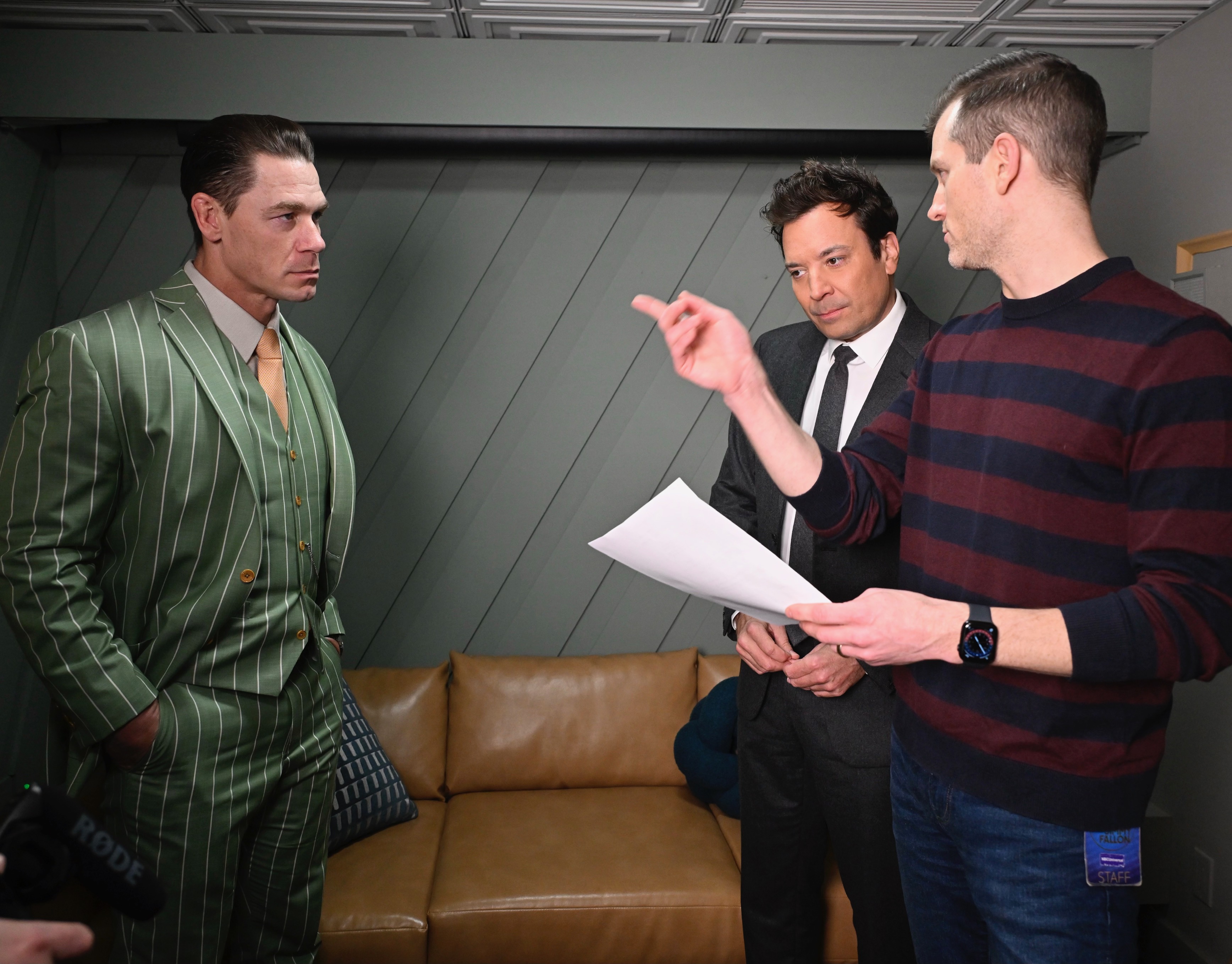 Nick Dyer ’11 holds a sheet of paper and gestures while speaking with John Cena and Jimmy Fallon in a backstage room.