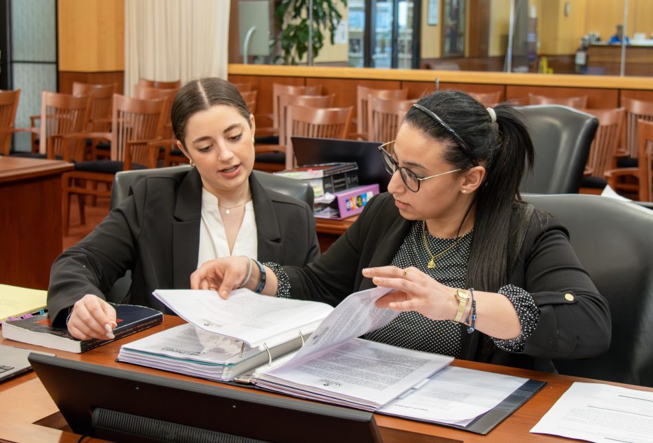 Two people seated at a desk in a courtroom. They are paging through binders of notes.