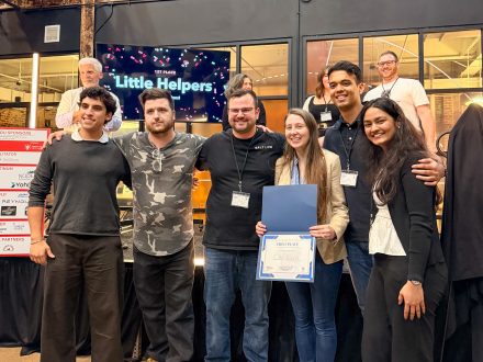 Six members of the Little Helper team pose together after receiving first place, with the first-place announcement displayed behind them.