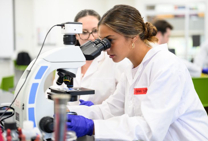 A student looks through a microscope during a microbiology lab as another student observes behind her.