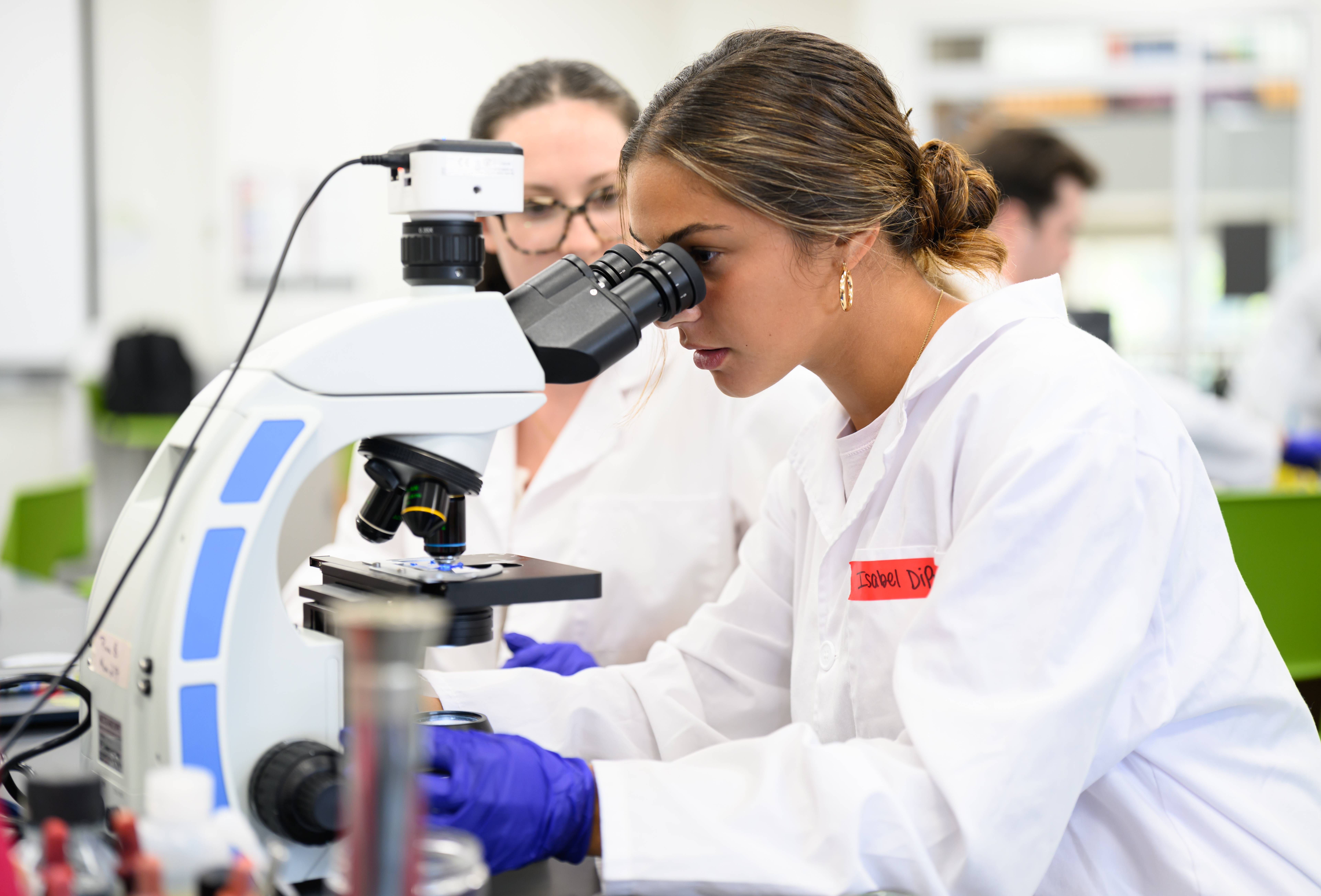 A student looks through a microscope during a microbiology lab as another student observes behind her.