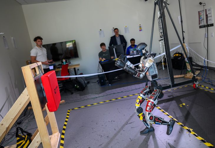 A humanoid robot moves inside a research lab as students and faculty watch from behind a rope.