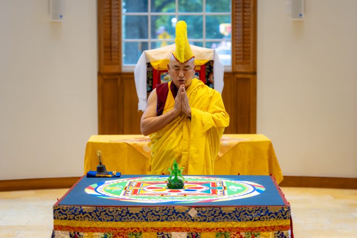 A Buddhist monk stands with his hands together in prayer behind a colorful sand mandala.