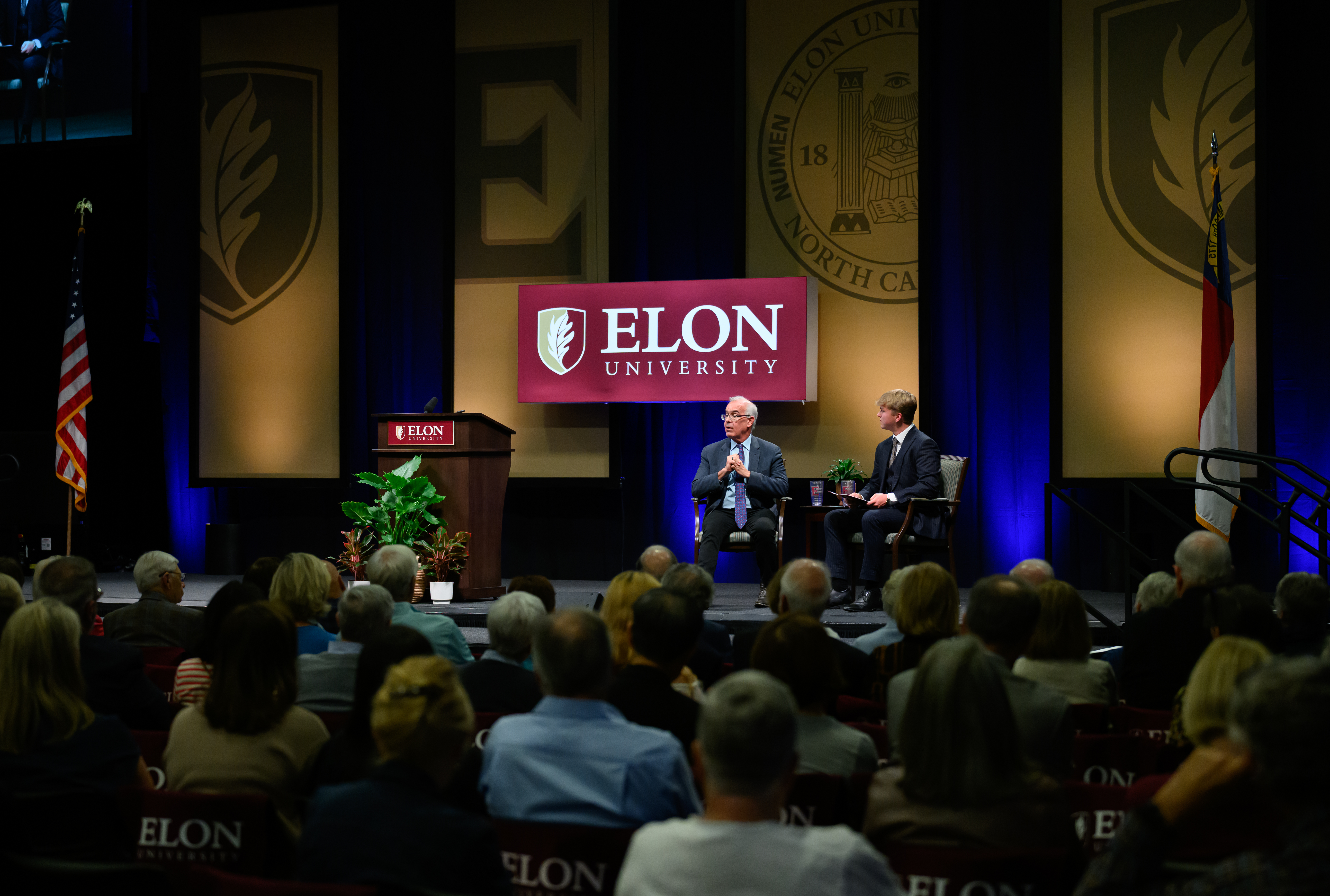 A speaker and moderator sit onstage in Alumni Gym during Elon’s Fall Convocation as audience members watch.