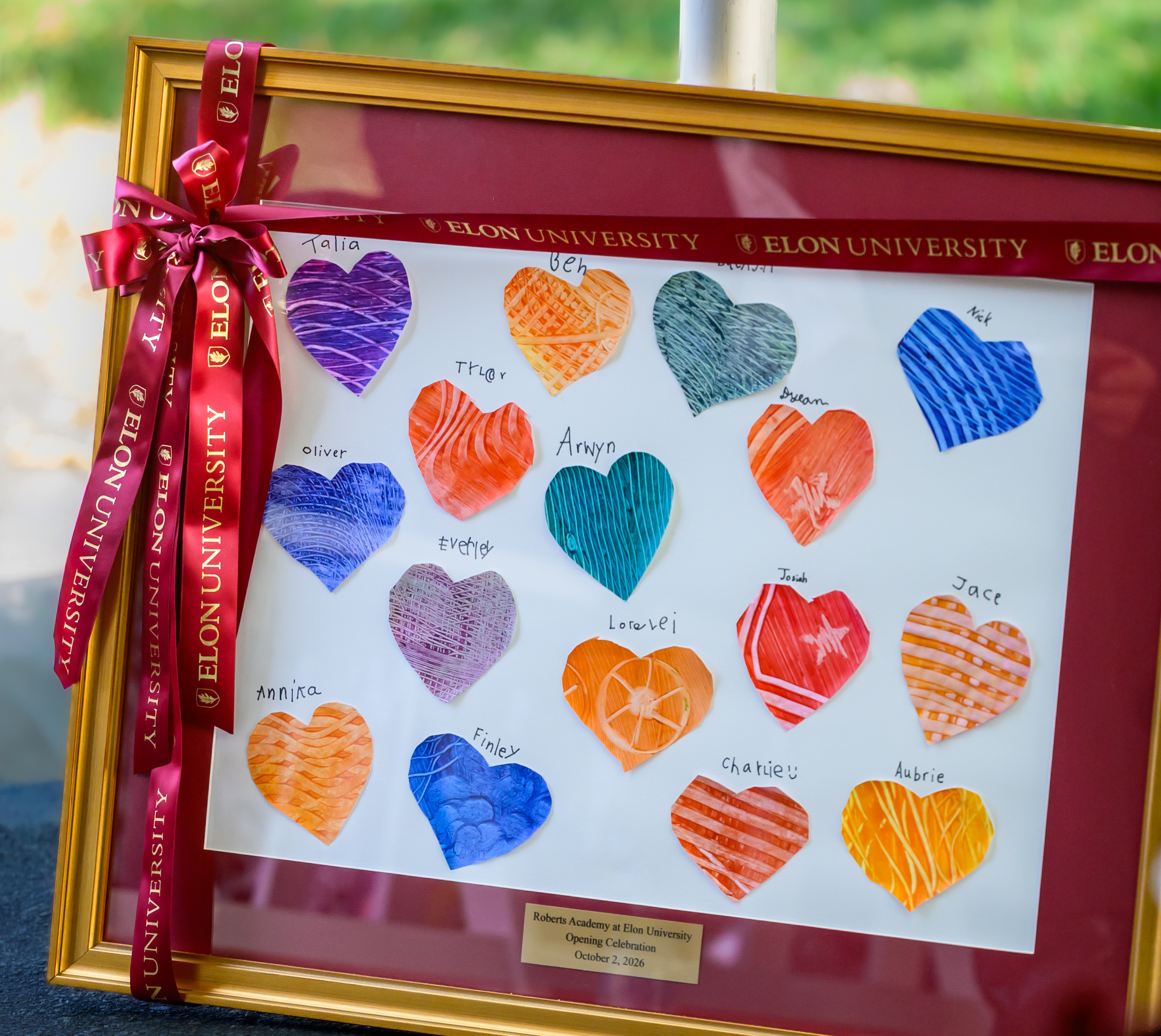 A photo of a framed piece of art that has paper hearts mounted on display with the names of each student enrolled in the charter class of the Roberts Academy at Elon University.