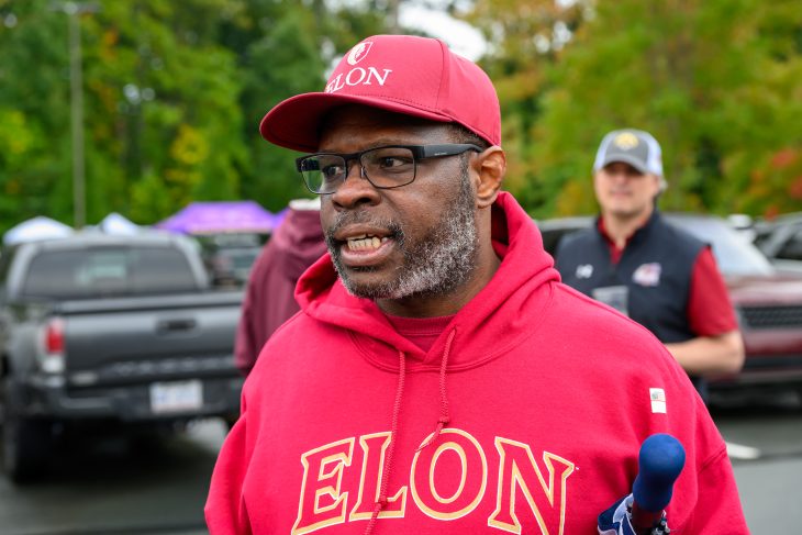 A man wearing a maroon Elon hoodie and matching baseball cap speaks into a microphone during Homecoming festivities.