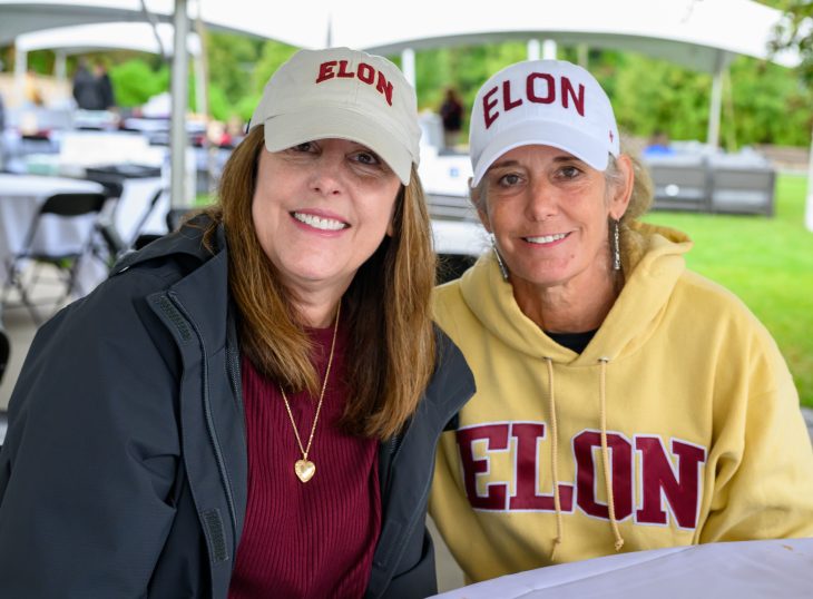 Two women wearing Elon baseball caps smile together beneath a tent during Homecoming Weekend.