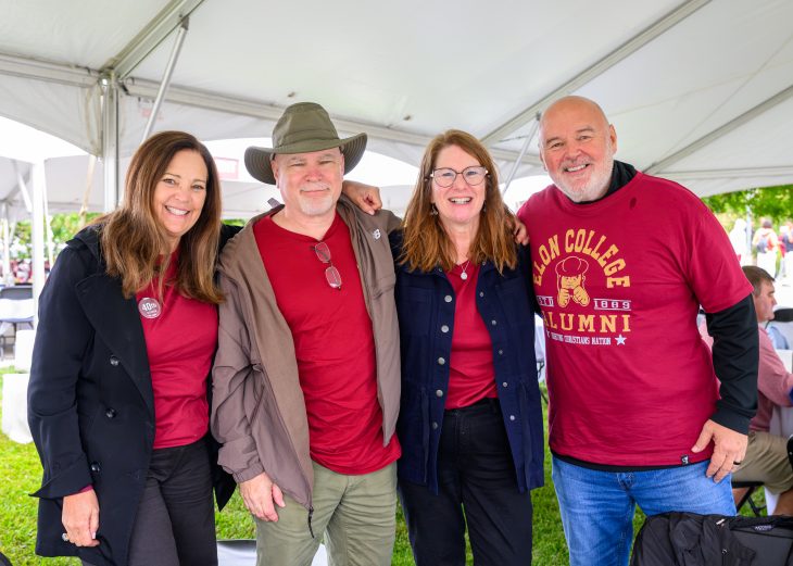 Four Elon alumni pose together beneath a white tent, wearing maroon shirts and jackets during Homecoming celebrations.