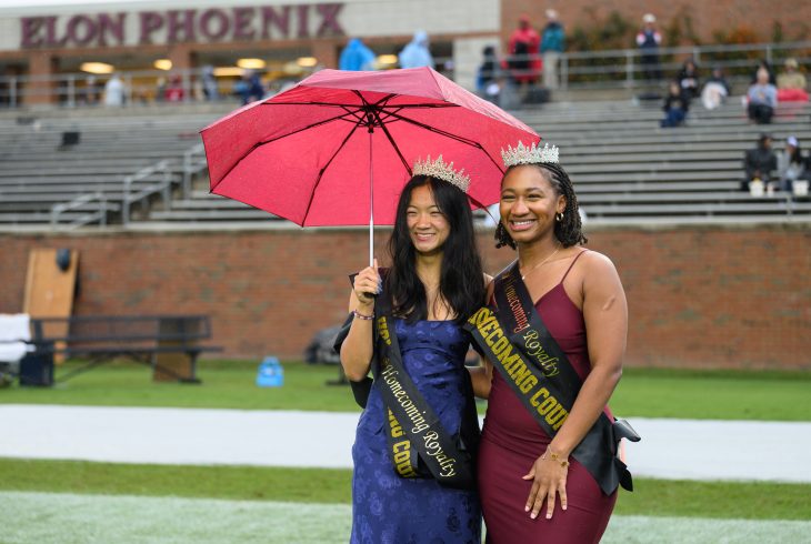 Two young women stand in dresses with Homecoming Royalty sashes. One holds an umbrella.