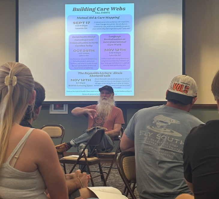 A photo of a man with a beard sitting in front of a room talking to people sitting