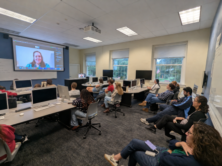 Students and faculty seated in a computer classroom in Alamance 318 watch PWR alumna Margaret Achey speak via Zoom on a large projection screen.