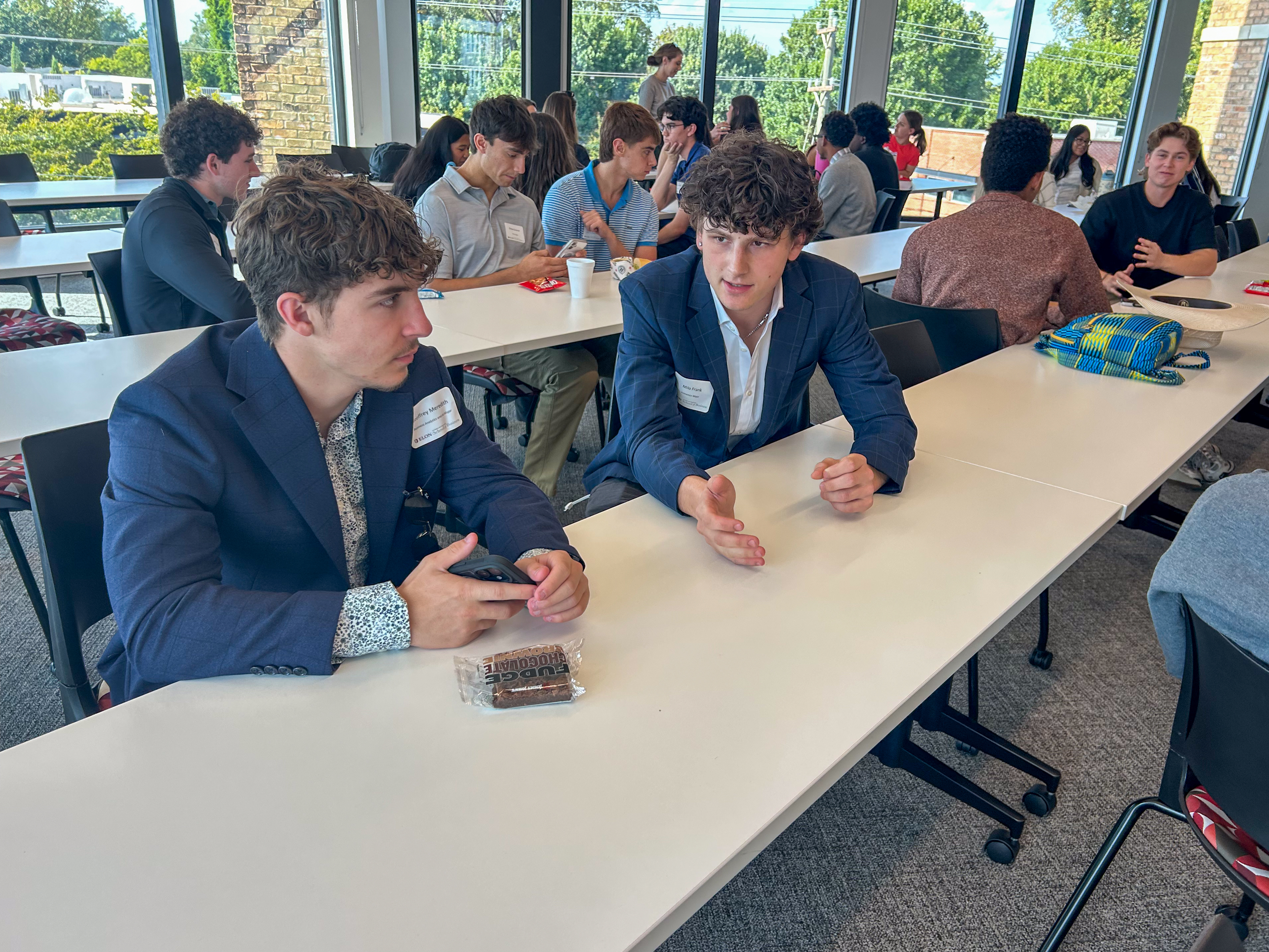 Two Elon students talk across a classroom table as other students converse in small groups behind them.