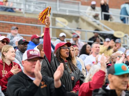 Elon fans cheer and clap in the stands at Rhodes Stadium, with one fan waving a maroon and gold pom-pom.
