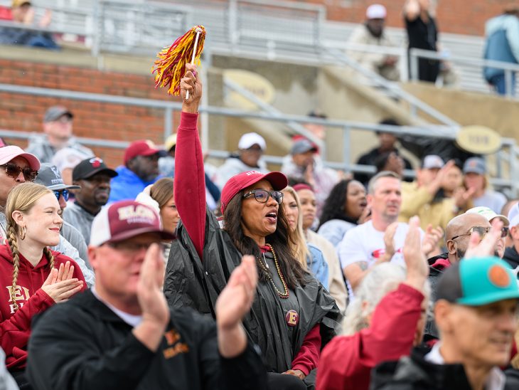 Elon fans cheer and clap in the stands at Rhodes Stadium, with one fan waving a maroon and gold pom-pom.