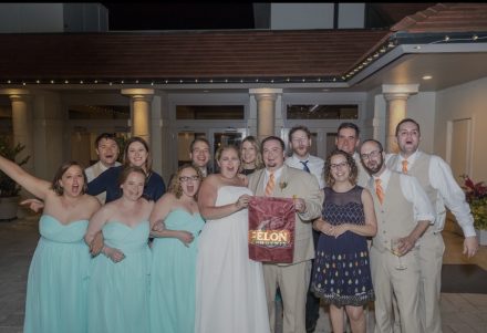 Kat and Chris Wallace pose with their wedding party and fellow Elon alumni while holding an Elon Phoenix banner.