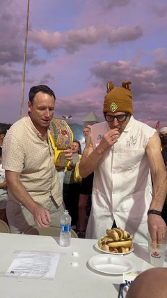 Joel Karty in a lab white coat eating hot dogs as Joey Chestnut stands next to him cheering him on during the hot dog eating contest.