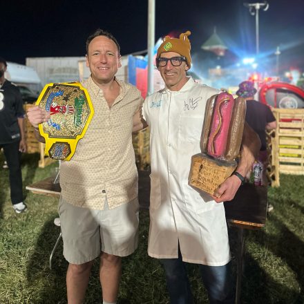 Joel Karty in a lab white coat holding a wood carved hot dog trophy standing next to Joey Chestnut whole is holding a belt for winning a hot dog eating contest.