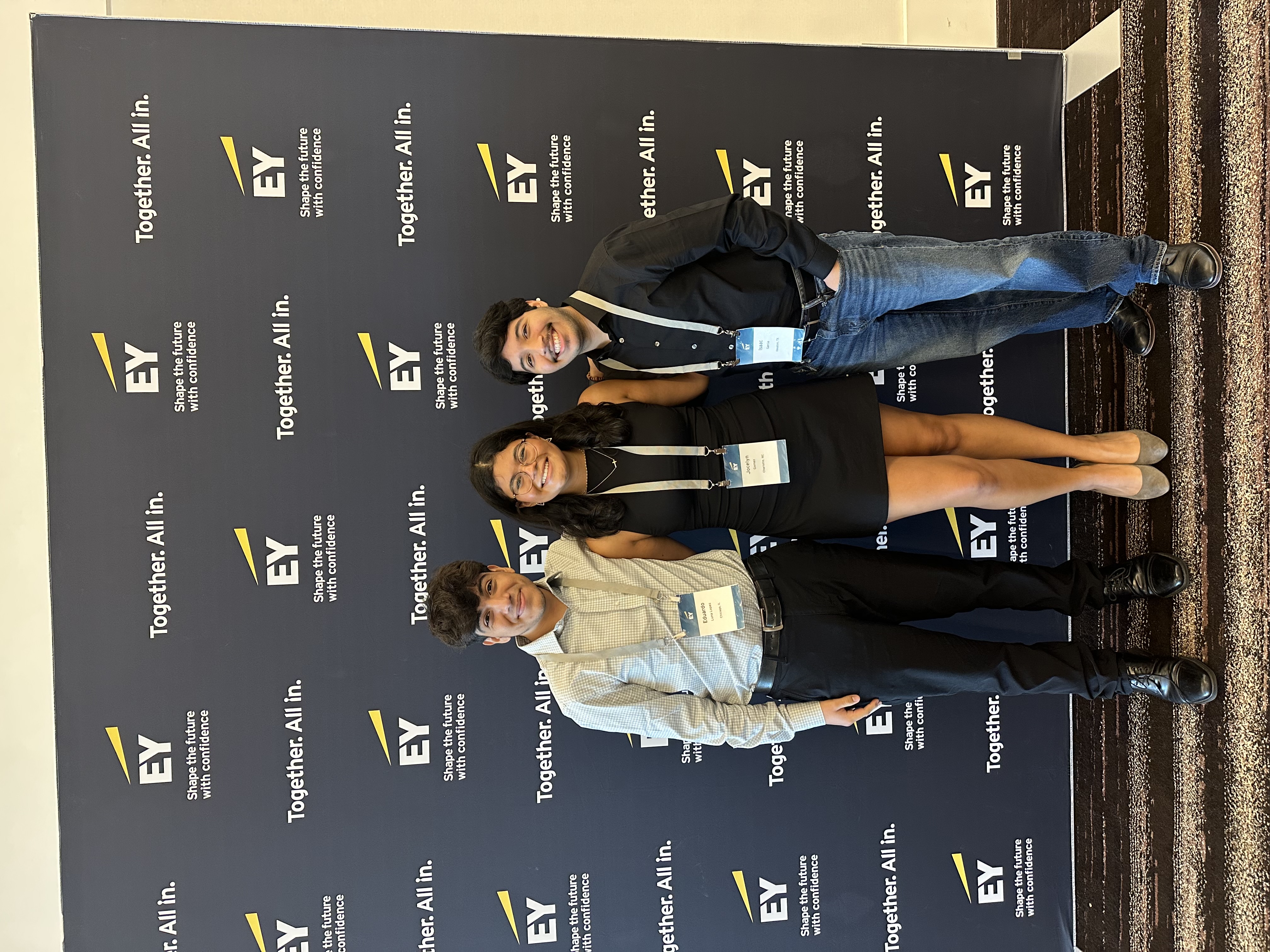 Jocelyn Gomez ’27 stands between two fellow attendees wearing name badges in front of an EY backdrop.