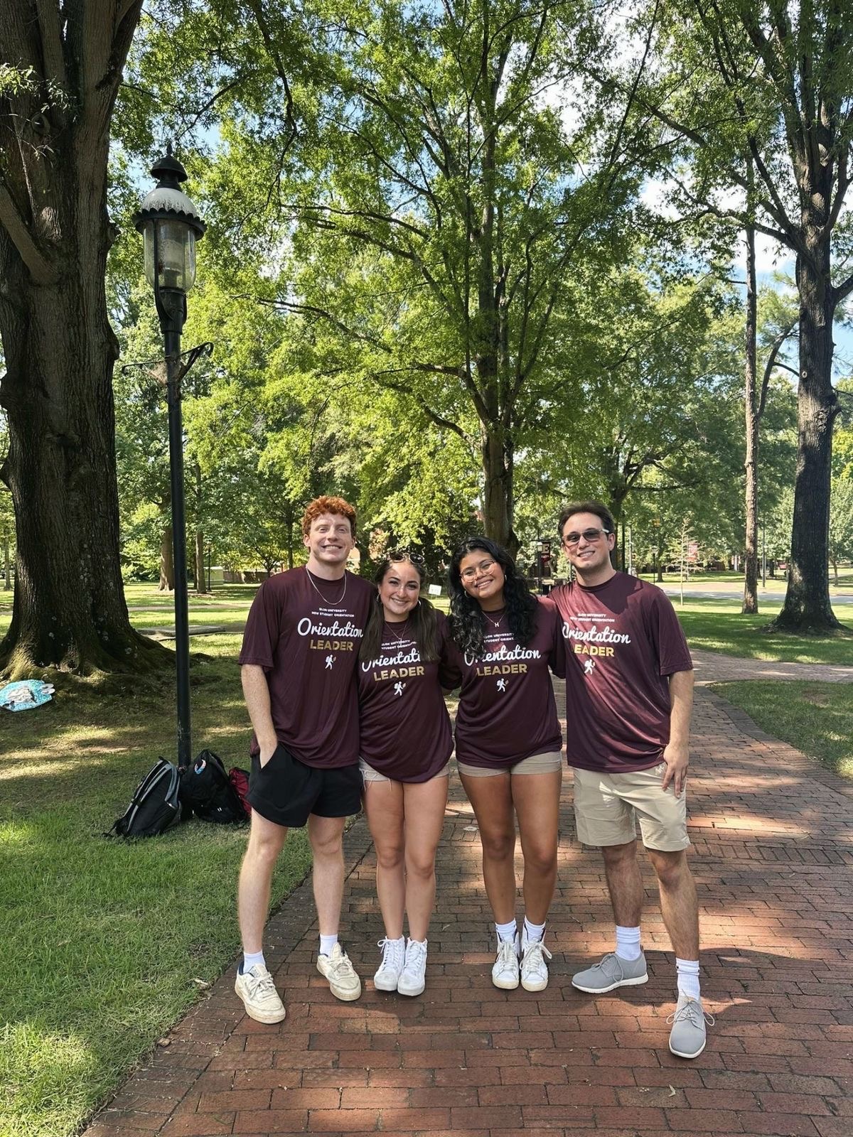 Four Elon orientation leaders in maroon shirts pose on a tree-lined brick path, with Jocelyn Gomez ’27 second from right.
