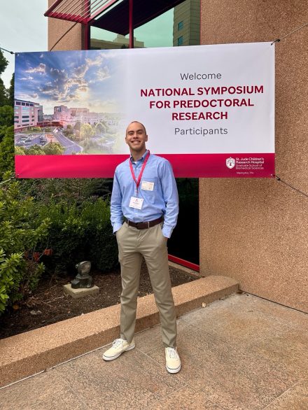 Alex Roberts is wearing a button down dress shirt and khakis while standing in front of a sign for the St. Jude Graduate School National Symposium for Predoctoral Research.