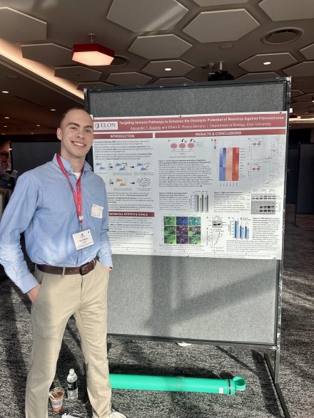 Alex Roberts is wearing a button down dress shirt and khakis while standing in front of his poster at the poster presentation.
