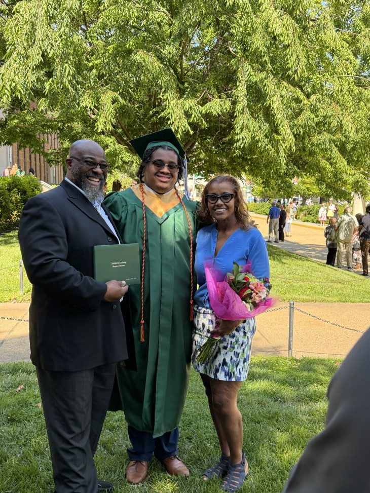 A graduate in a green cap and gown stands between two adults outdoors.