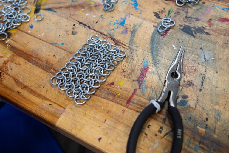 Chainmail on a table next to a bent-nose pliers