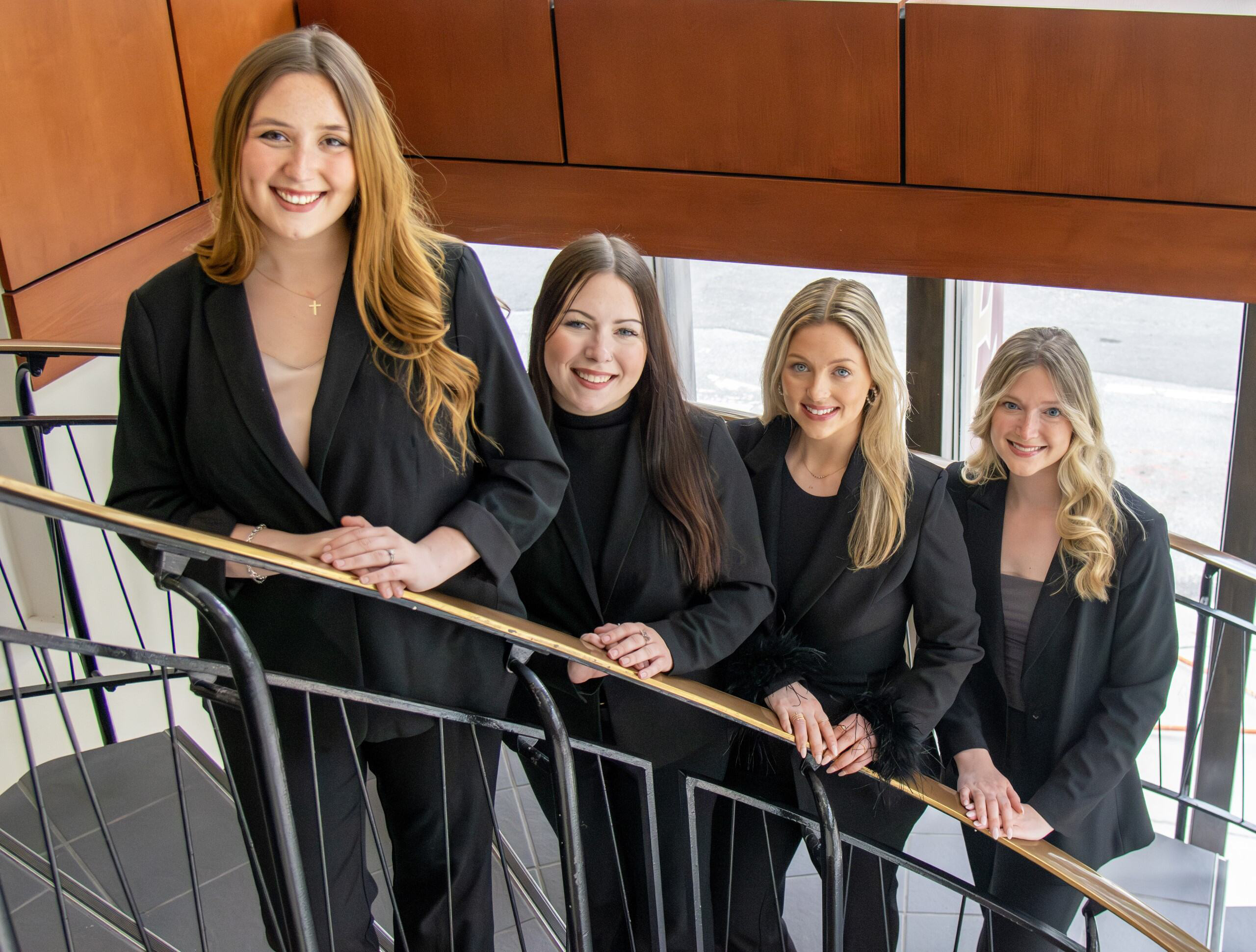 Four women law students posed on a spiral staircase. They are wearing black suits.