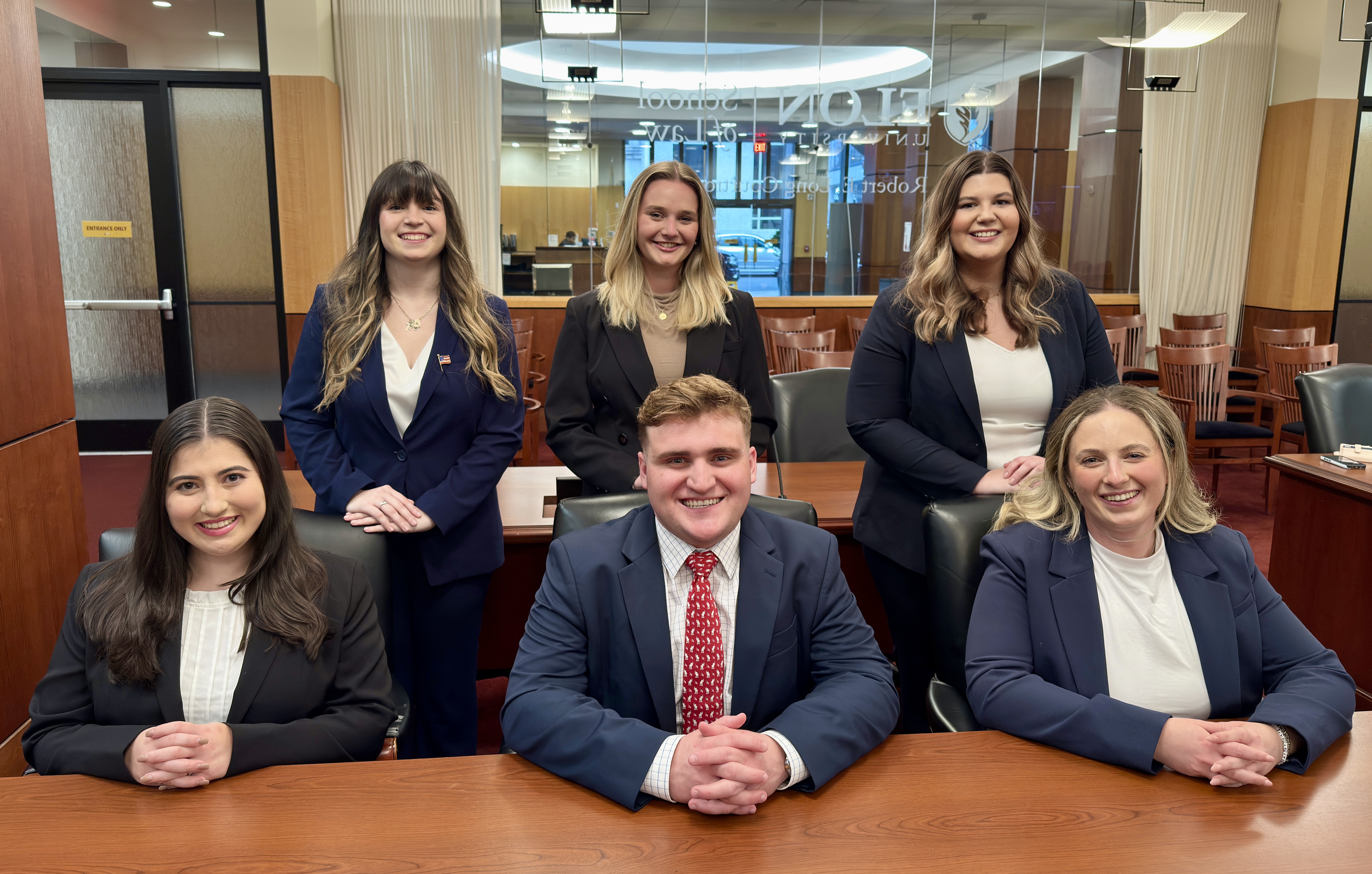 A group of students seated at and standing around a desk in the Robert E. Long Courtroom at Elon Law