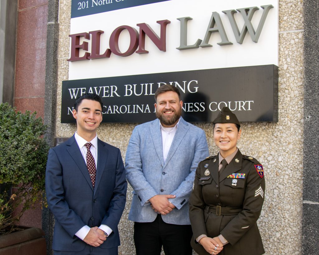 Three law students in military dress and professional attire in front of the Elon Law - Weaver Street Building sign on North Greene Street.